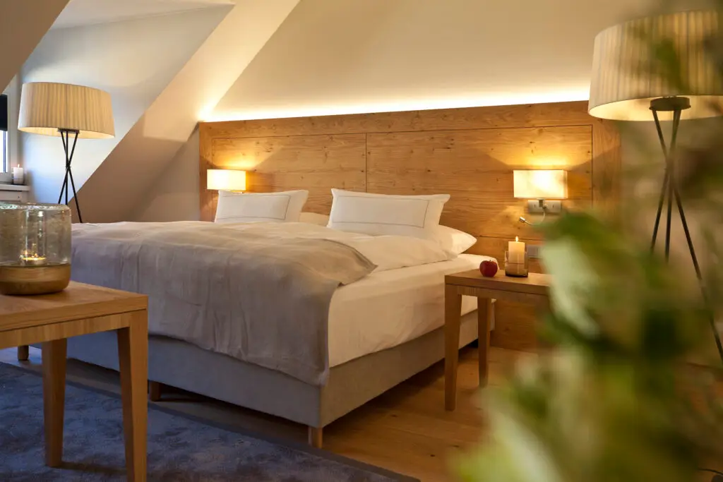 Großes Doppelzimmer mit elegantem Interieur und Doppelbett, darauf zwei Kopfkissen und hochwertige Bettwäsche. Rechts und links eingeschaltete Nachtlampen mit angenehmer und augenfreundlicher Beleuchtung. Rechts auf dem Nachttisch eine angezündete Kerze und ein roter Apfel. Im Vordergrund das verwischte Grün eines Blumenstraußes.