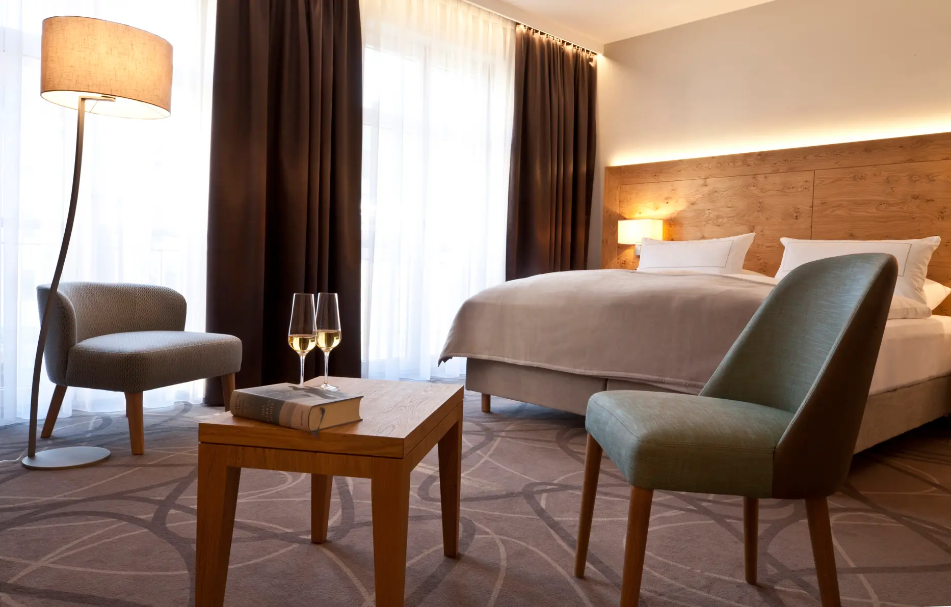 Hier sieht man eines der großen Doppelzimmer im Gasthof Zufriedenheit. Im Vordergrund einladende kleine Sessel mit einem kleinen Tisch in der Mitte, darauf ein Buch und zwei gefüllte Sektgläser. Dahinter dann das Kingsize-Bett mit Tagesdecke. Es dominieren die Farben helles Salbei und warme Naturholztöne. Das Zimmer sieht geräumig und sehr wohnlich aus. Ein großzügiges en-suite Badezimmer, moderne Flatscreen TVs und WIFI sind genauso selbstverständlich wie feine natürliche Stoffe und leichte Daunendecken.