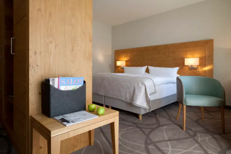 Auf diesem Bild ist eines der Gartenzimmer zu sehen, mit einem Kingsize Bett, das Kopfteil aus heller Roseneiche. Auf dem Boden Auslegware, die eingeschalteten Nachttischlampen verbreiten behagliches Licht. Vor dem Bett steht ein kleiner Sessel mit salbeifarbenem Bezugsstoff. Auf einem kleinen Sofatisch steht ein Zeitschriftensammler mit Magazinen und es liegen zwei frische, knackige grüne Apfel auf dem Tisch.