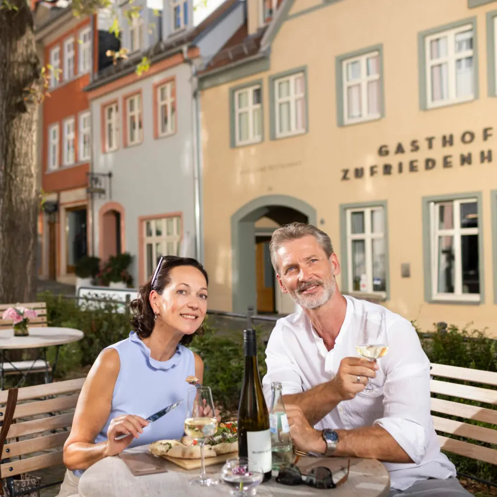 Es sitzt ein attraktives Pärchen mittleren Alters auf der Freifläche vor dem Gasthof Zufriedenheit auf den hellen Holzmöbeln. Sie haben beide etwas zu Essen vor sich auf ihren Tellern und je ein Glas Weißwein.. Er hält sein Glas in der Hand und schaut Richtung UNESCO Welterbe Naumburger Dom und beide zusammen machen den Eindruck, als würden sie während des Essens angeregt Pläne schmieden über die nächsten Tage. Das Licht-Schattenspiel vermittelt den Eindruck eines warmen Sommertages.