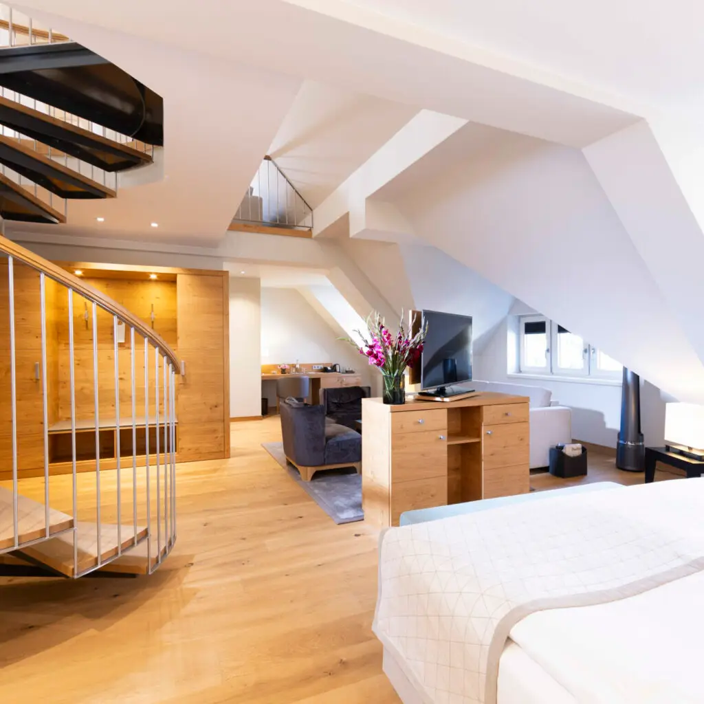Zu sehen ist die große Maisonette-Suite, vorne links sieht man die Wendeltreppe, die nach oben führt und ebenfalls im Vordergrund das einladende Doppelbett mit hochwertiger weißer Bettwäsche. Weiter hinten im Bild dann die punktuell ausgeleuchtete Garderobe, ein Sideboard, was als Raumteiler dient, darauf der schwenkbare Fernseher und ein großer Strauß frischer Blumen. Rechts im Foto sind die Fenster zu erkennen, die auf die Fußgängerzone Steinweg führen, der wiederum in 2 Gehminuten zum UNESCO Welterbe Naumburger Dom führt. Hinter dem Sideboard aus hochwertigem, hellen Holz schaut man auf die anheimelnde Sitzgruppe. Diese Maisonette Suite ist insgesamt rund 60qm groß.