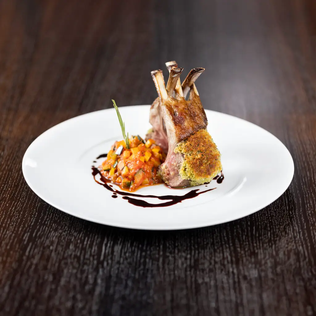 Flacher, edler, weißer Porzellanteller auf dunklem Holztisch mit kunstvoll angerichtetem Lammkarree, überzogen mit einer knusprigen Kräuterkruste. Dazu ein Ratatouille Gemüse mit einem Zweiglein Thymian obenauf, und ein dezenter Spiegel vom dunklem Bratenjus.