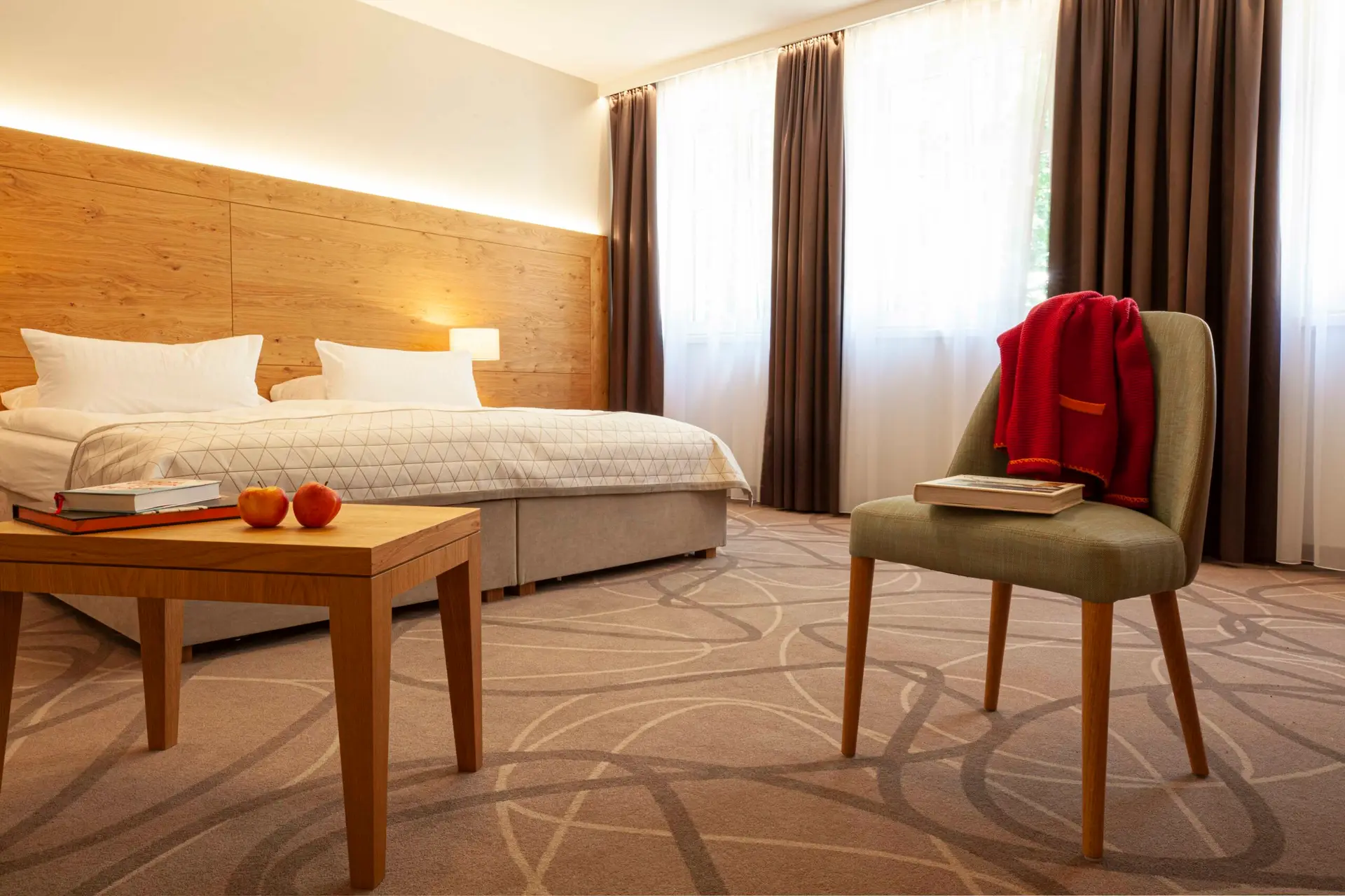 Hier ist das behindertenfreundliche Zimmer im kleinen, feinen Boutique Hotel Gasthof Zufriedenheit in Naumburg zu sehen. Es ist besonders geräumig; man sieht das breite Kingsize Bett und davor einen salbeifarbenen Stuhl mit einem Buch und einer orangefarbenen Jacke darübergelegt. Auf dem kleinen hellen Holztisch davor liegen zwei rote Äpfel und weitere Lektüre. Es entsteht der Eindruck von großzügiger Entspanntheit und einem wirklich geruhsamen Aufenthalt. Das Kopfende des Bettes ist aus heller, sandgestrahlter Roseneiche, was nicht rustikal, sondern elegant aussieht.