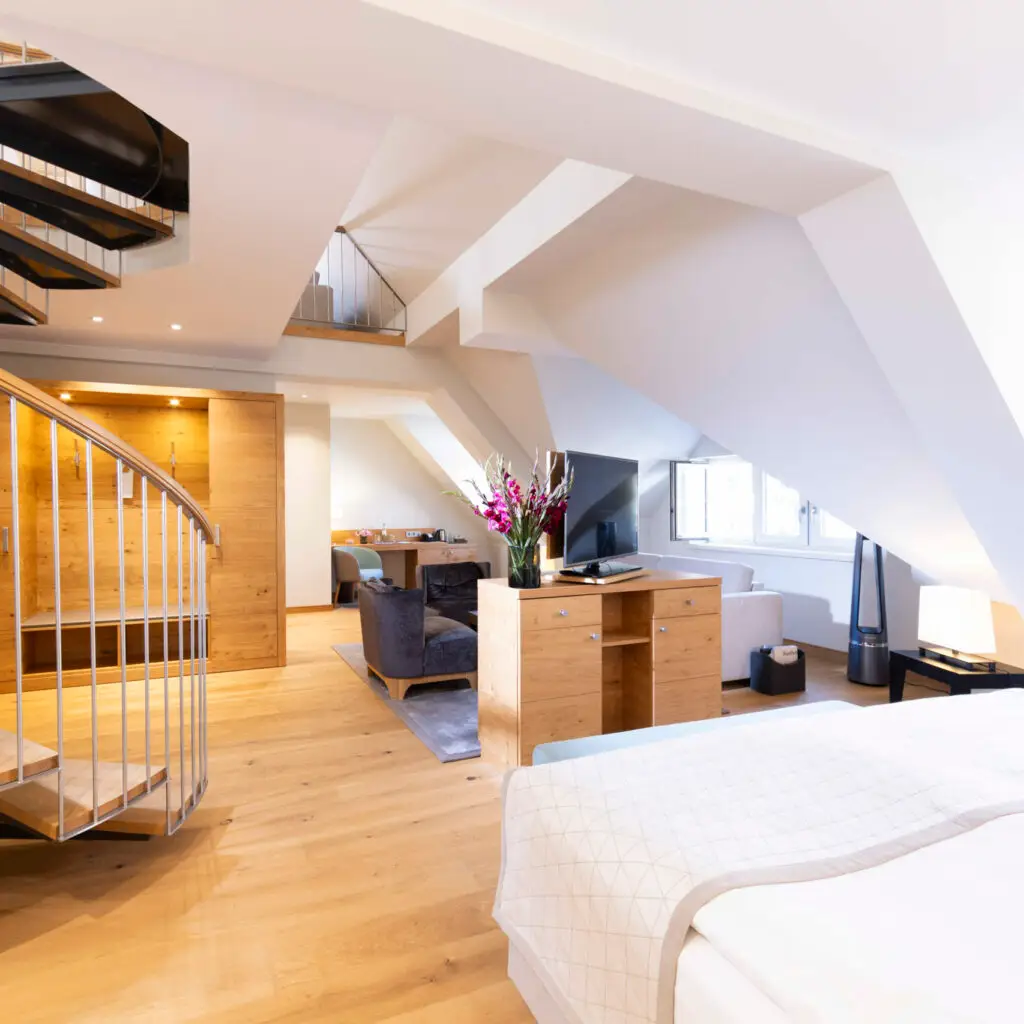 Zu sehen ist die große Maisonette-Suite, vorne links sieht man die Wendeltreppe, die nach oben führt und ebenfalls im Vordergrund das einladende Doppelbett mit hochwertiger weißer Bettwäsche. Weiter hinten im Bild dann die punktuell ausgeleuchtete Garderobe, ein Sideboard, was als Raumteiler dient, darauf der schwenkbare Fernseher und ein großer Strauß frischer Blumen. Rechts im Foto sind die Fenster zu erkennen, die auf die Fußgängerzone Steinweg führen, der wiederum in 2 Gehminuten zum UNESCO Welterbe Naumburger Dom führt (Keywords ;- )) Hinter dem Sideboard aus hochwertigem, hellen Holz schaut man auf die anheimelnde Sitzgruppe. Diese Maisonette Suite ist insgesamt rund 60qm groß.