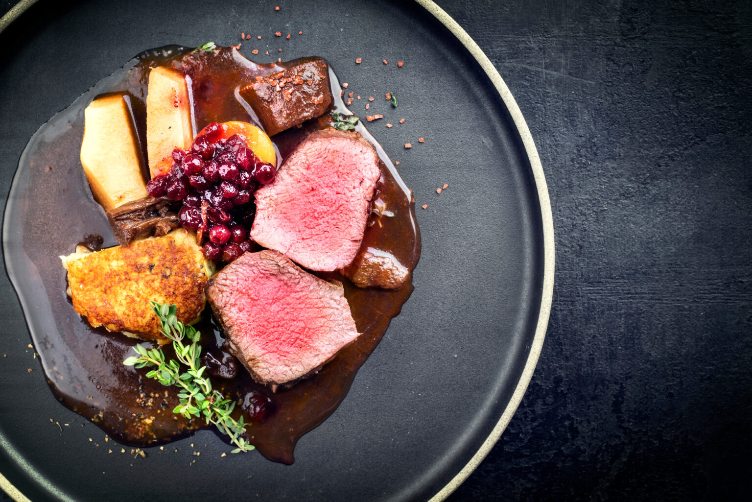 Traditional saddle of venison with fried mashed potatoes and fruits in game red wine sauce as top view on a modern design plate with copy space right